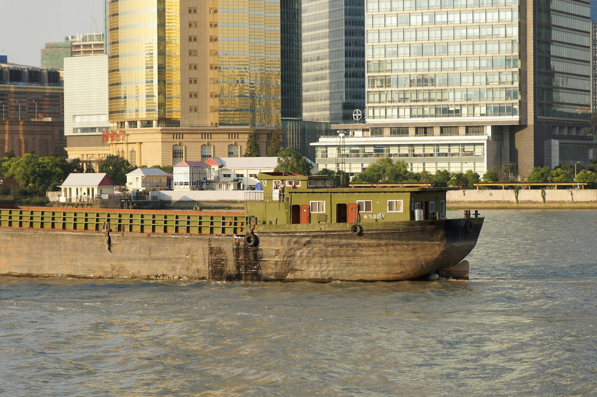 A cargo ship travels on a river, set against an urban skyline of modern office buildings.