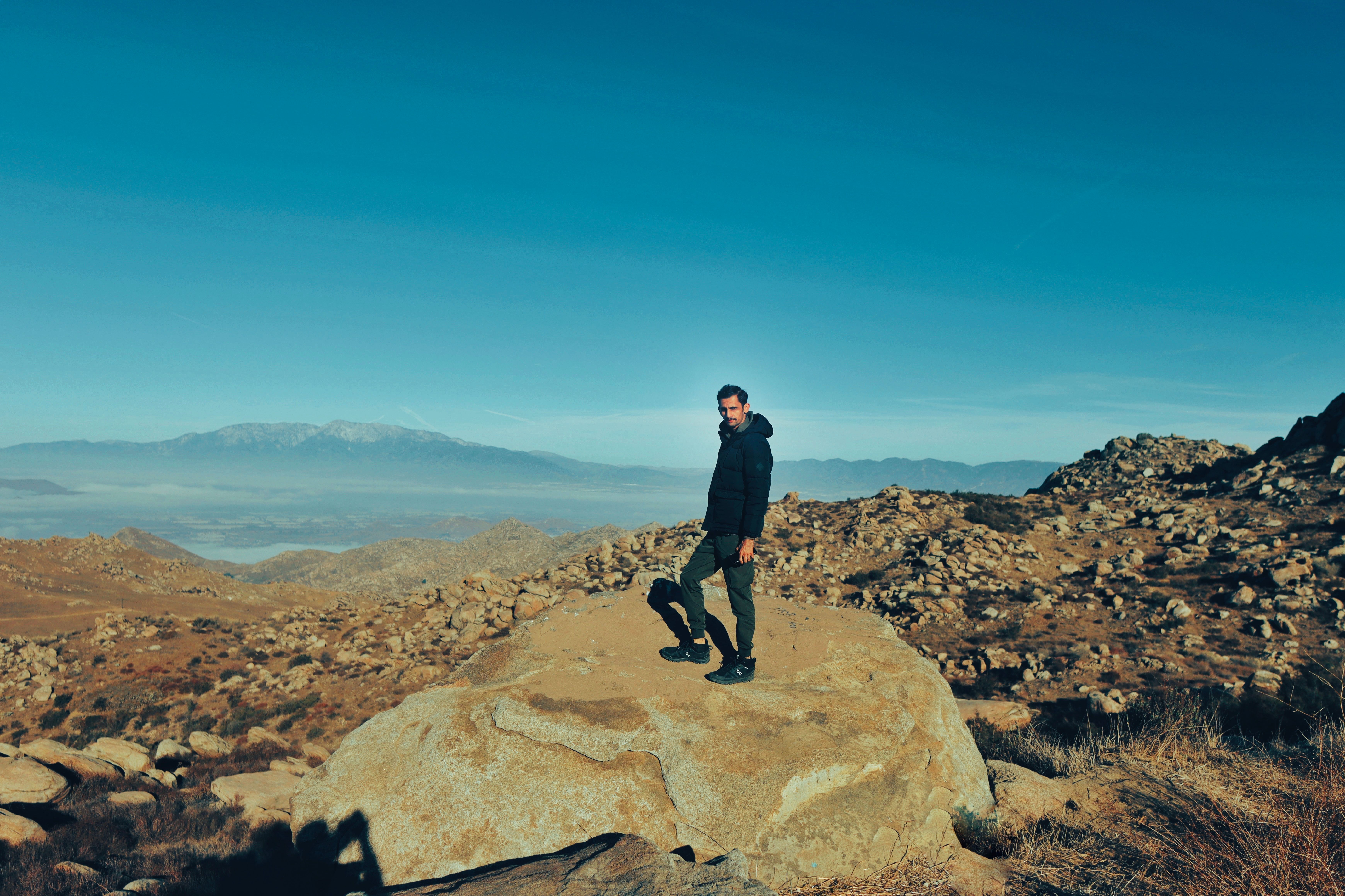 A man stands on a rock amidst a vast mountainous landscape under a clear blue sky.
