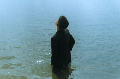 Free Rear view of a man in a suit standing in tranquil water, looking up contemplatively with a serene backdrop. Stock Photo