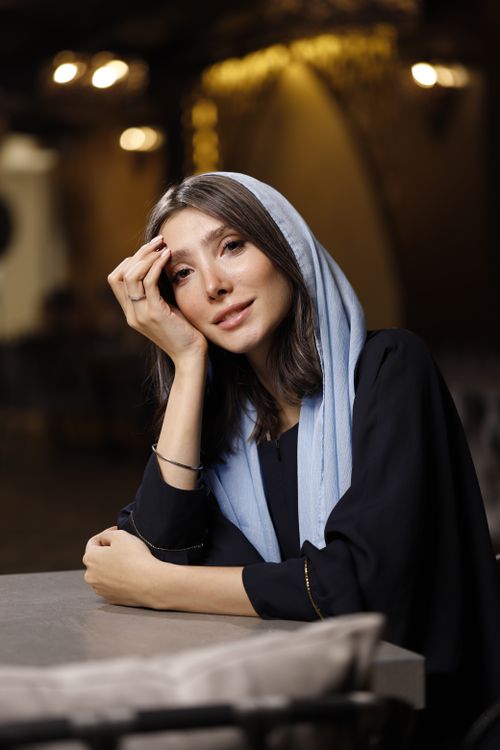 Free Portrait of a smiling young woman sitting indoors, wearing a light blue scarf. Stock Photo