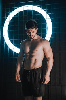 A shirtless man stands under a neon light circle, showing a fit, muscular physique.