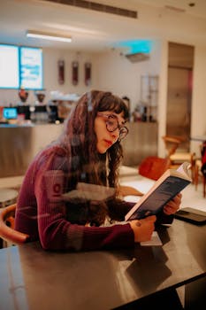 A young woman in a cozy café, absorbed in a book by a window. Captures a moment of relaxation and focus.