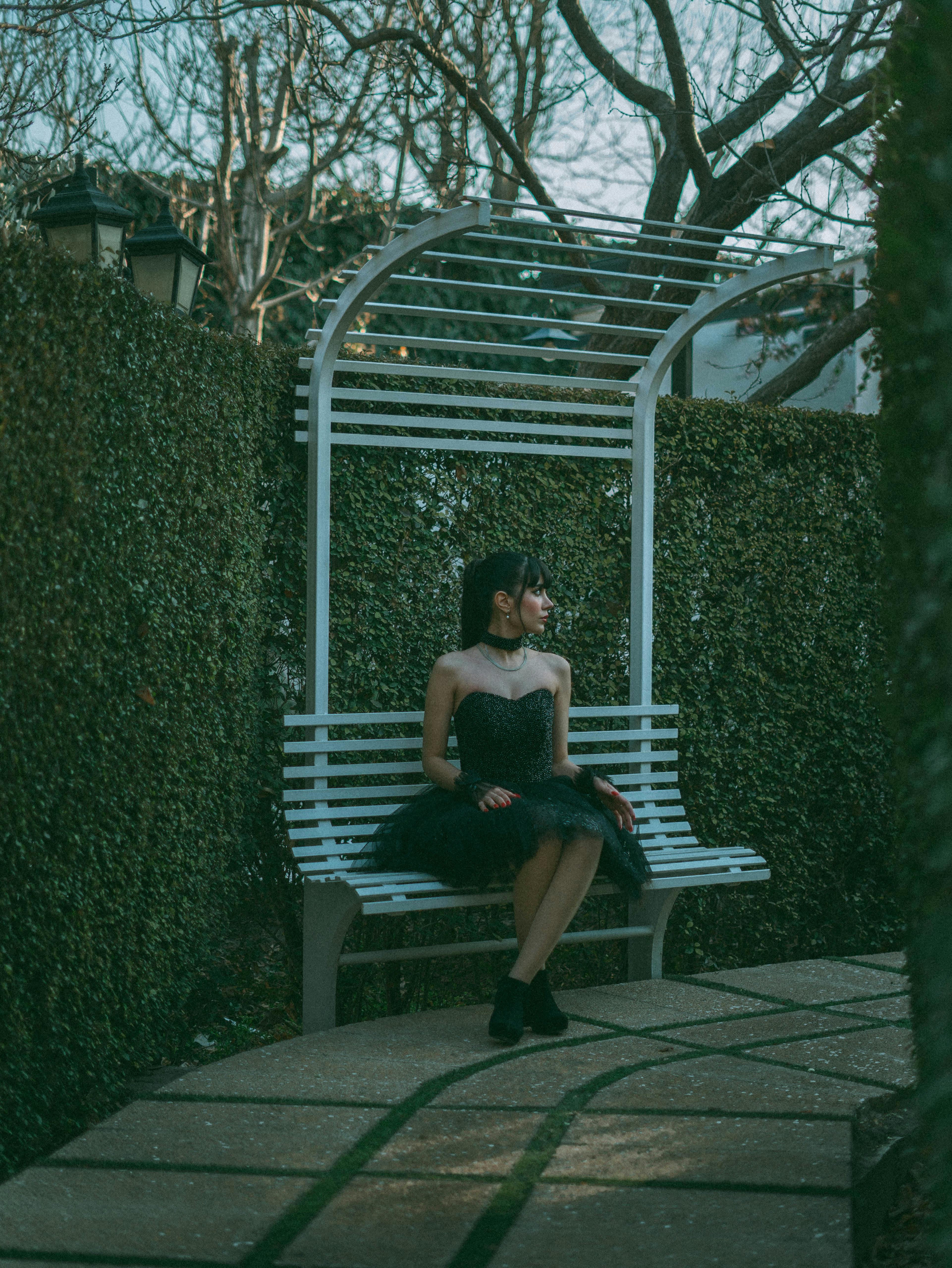 A woman in a black dress sits on a bench surrounded by lush garden foliage.