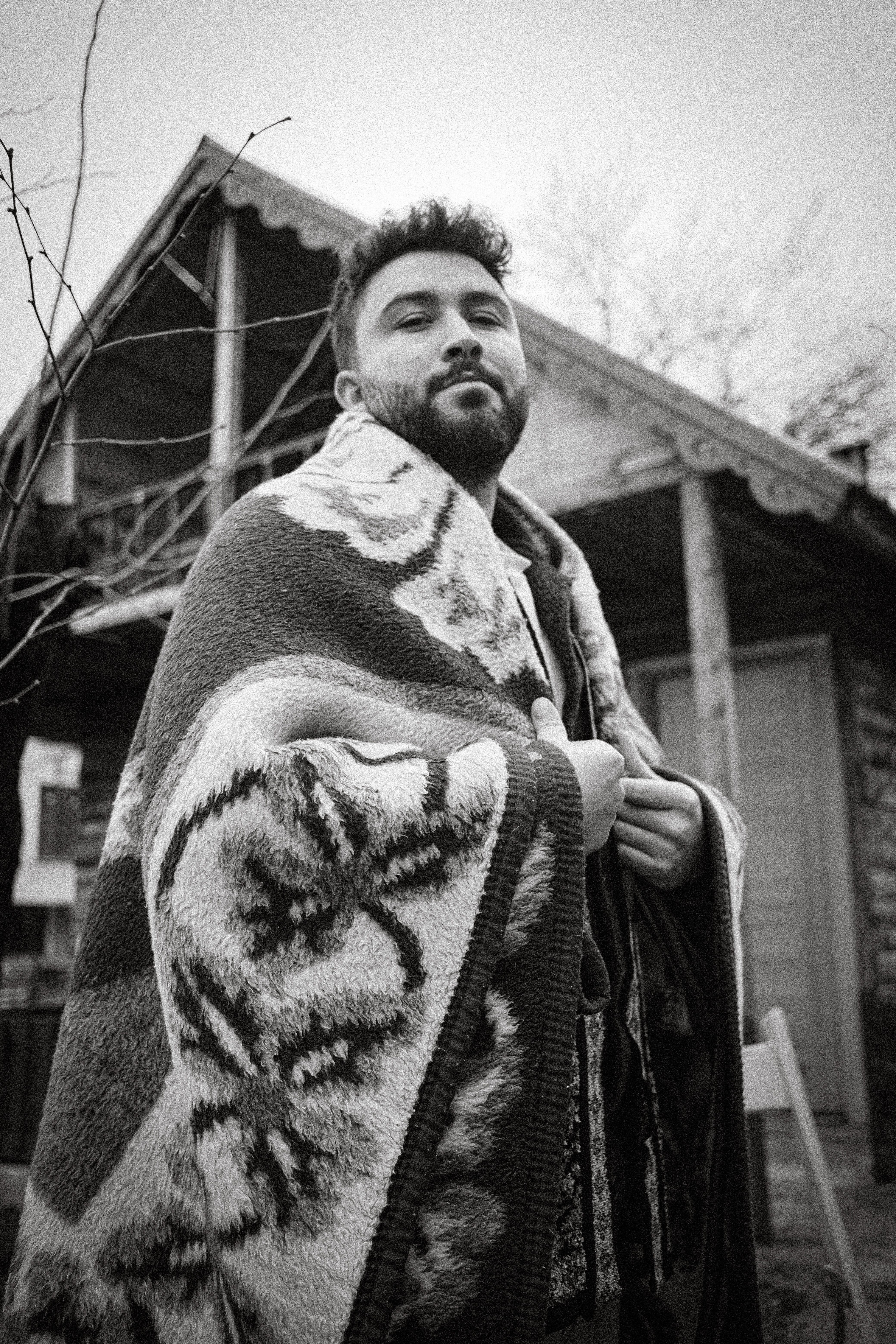 Man wrapped in a patterned blanket in front of a wooden cabin.