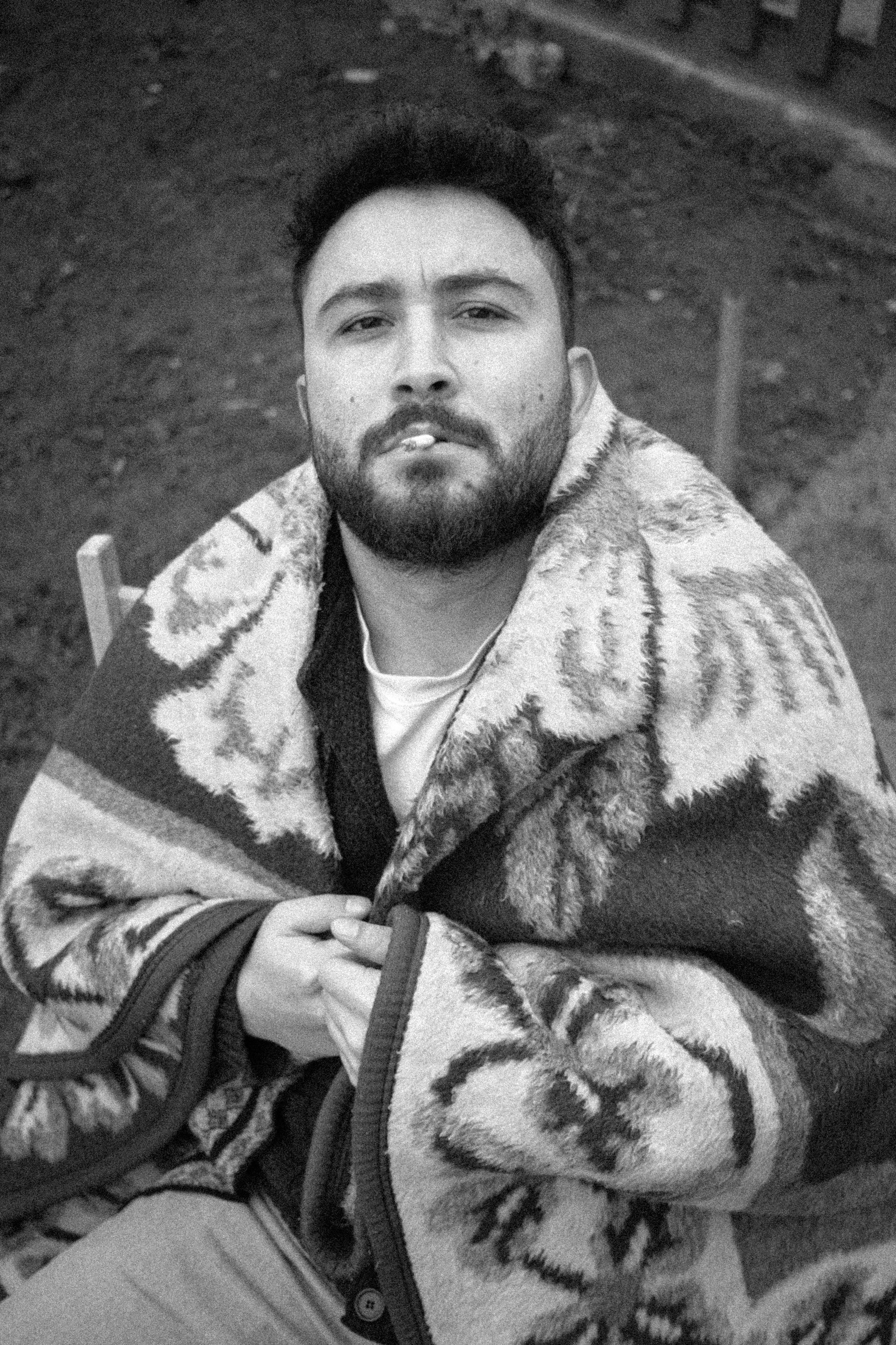 Bearded man wrapped in blanket, outdoors, thoughtful expression, black and white.