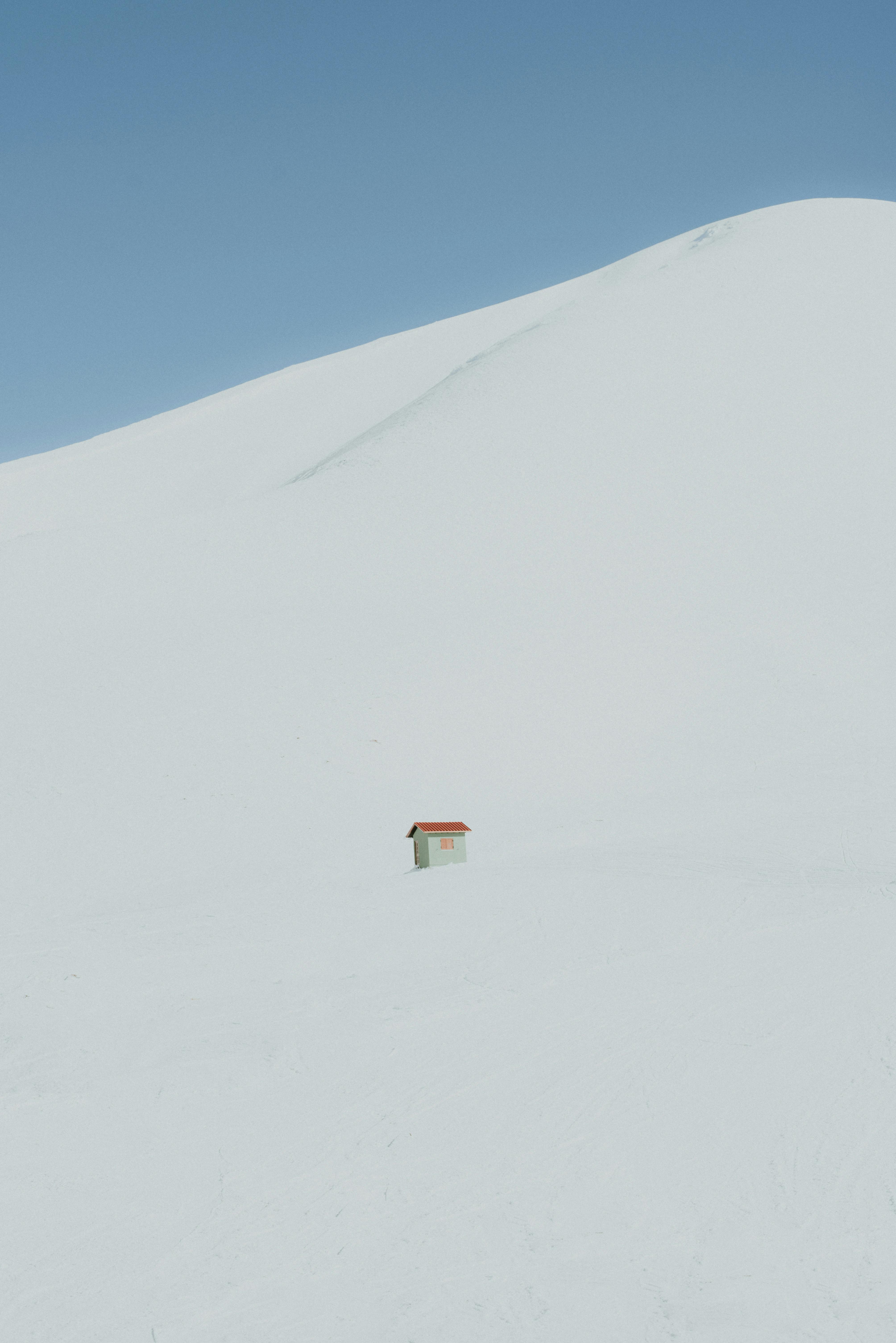 A small cabin stands isolated in a vast, snow-covered landscape under a clear blue sky.