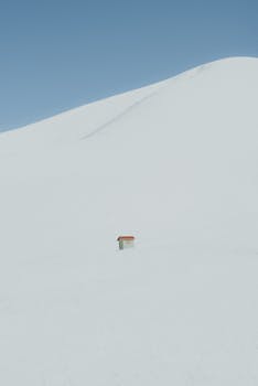 A small cabin stands isolated in a vast, snow-covered landscape under a clear blue sky.