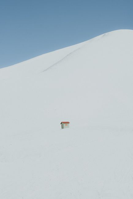 A small cabin stands isolated in a vast, snow-covered landscape under a clear blue sky.