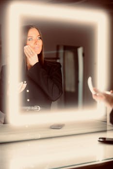 Woman in black jacket applying makeup in front of a square illuminated mirror indoors.