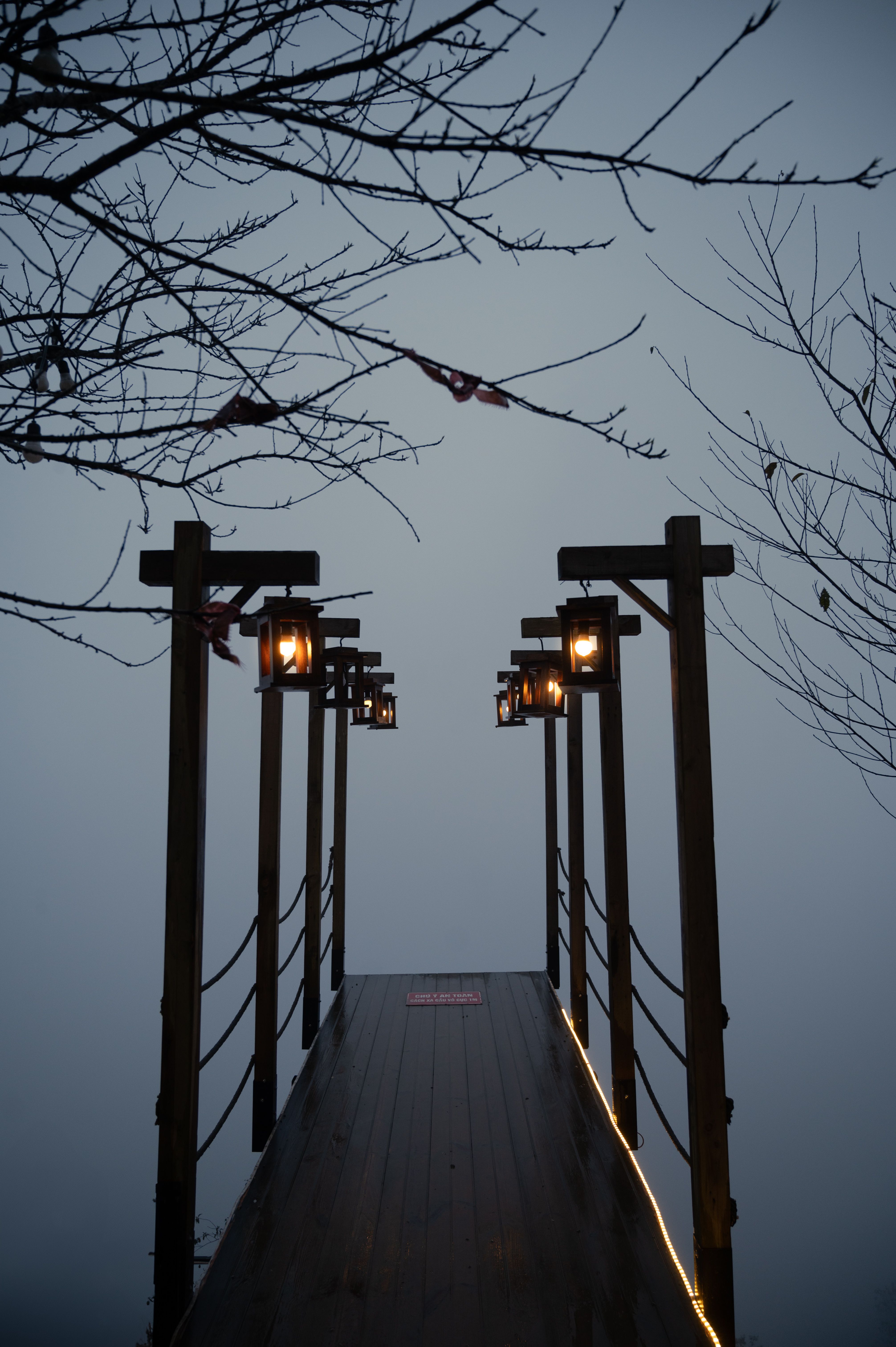 Wooden bridge lit by lanterns against a misty evening sky, photo by Nguyen Ngoc Tien on Pexels