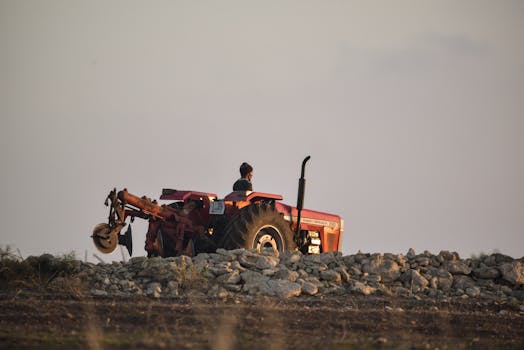 A lone farmer drives a red tractor over rocky terrain under a soft morning light.