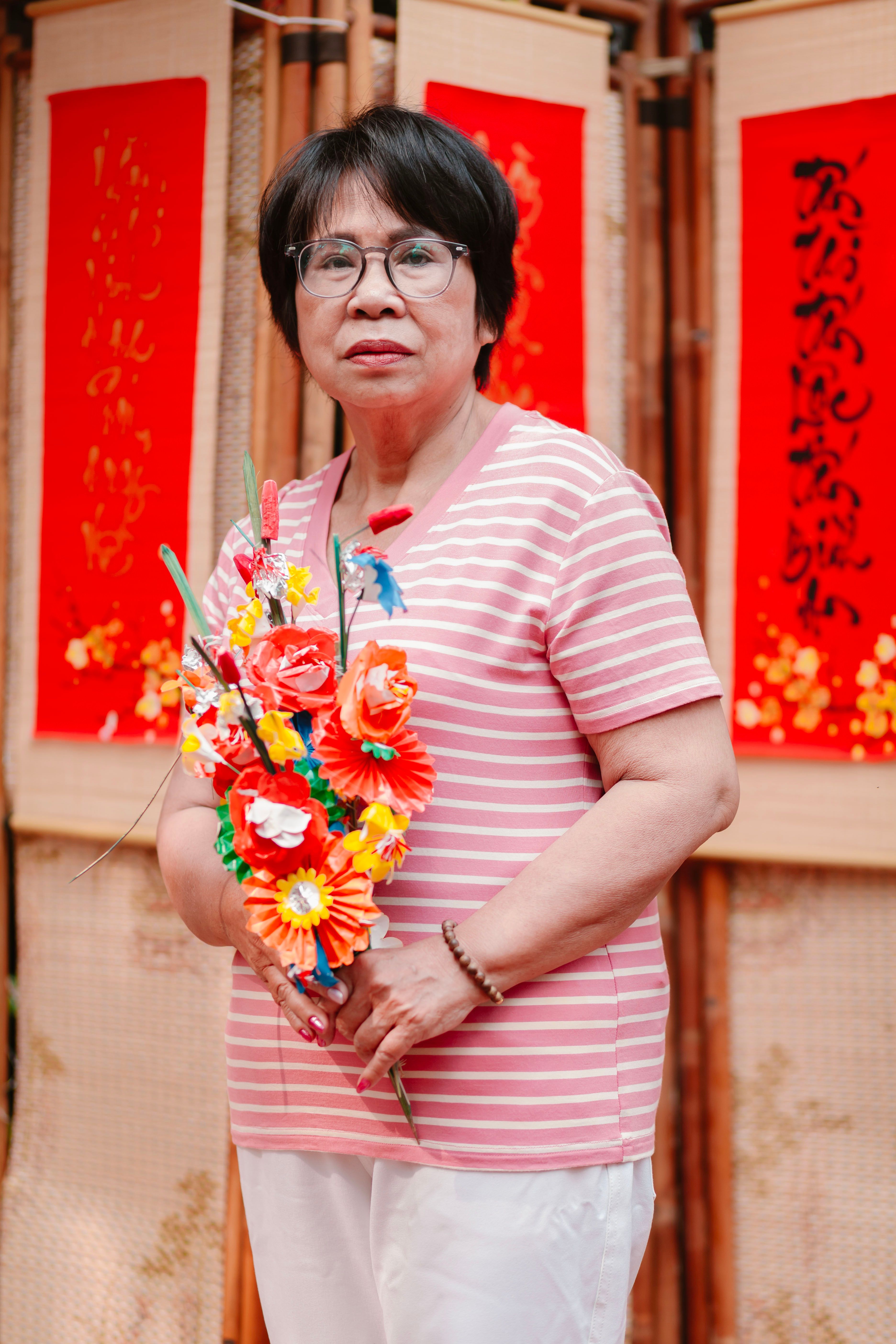 Elderly woman with paper flowers, standing in front of red Chinese scrolls.