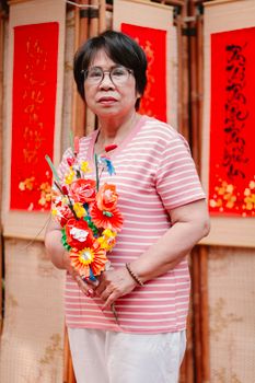 Elderly woman with paper flowers, standing in front of red Chinese scrolls.