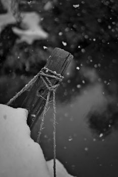 A snow-covered wooden post stands by a serene winter pond with falling snowflakes.