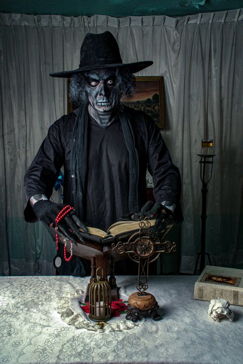 Free A costumed figure with a skeletal face reads a ritual book surrounded by gothic decor indoors. Stock Photo