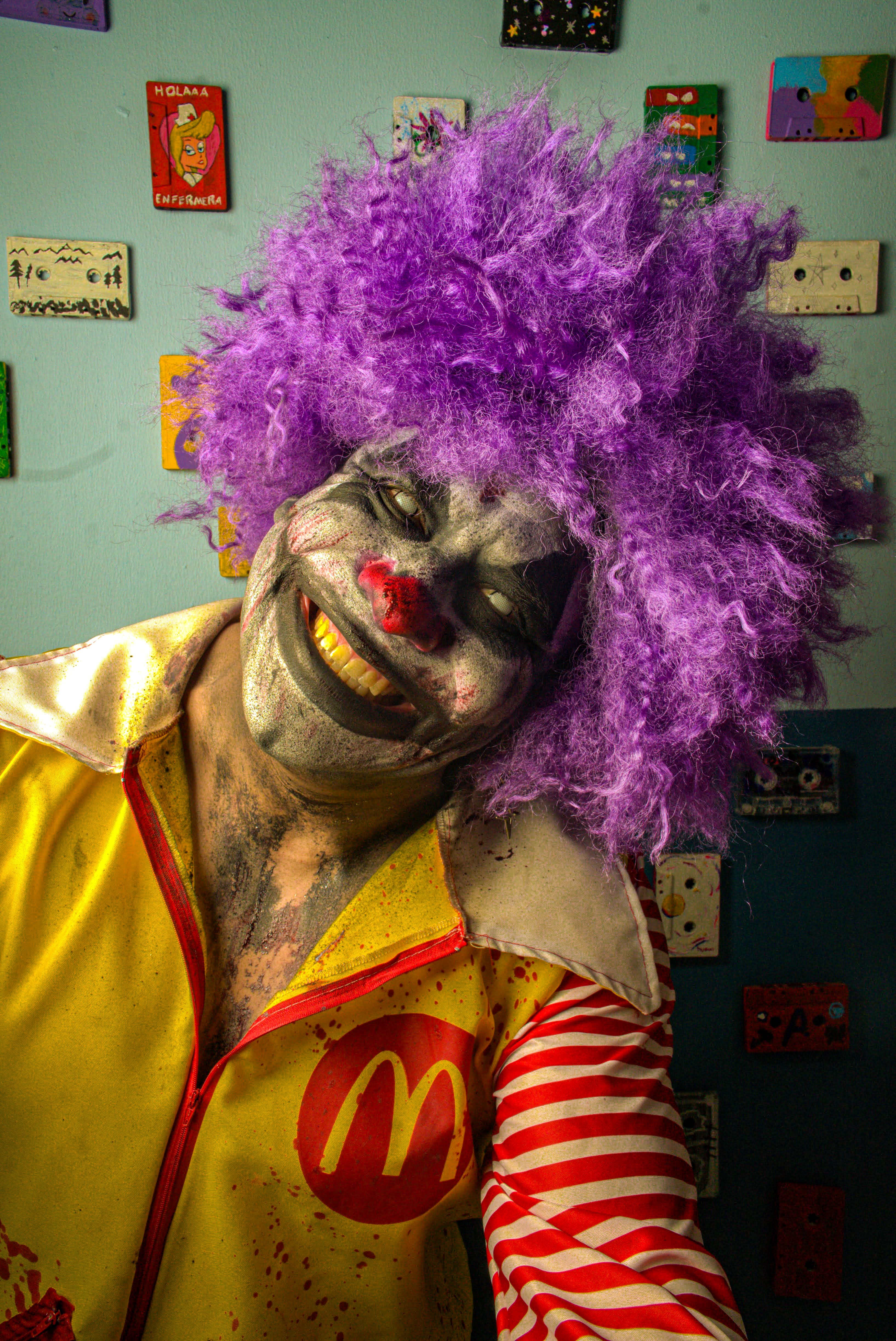 Colorful clown with purple wig, vivid costume, smiling mischievously inside a decorated room.