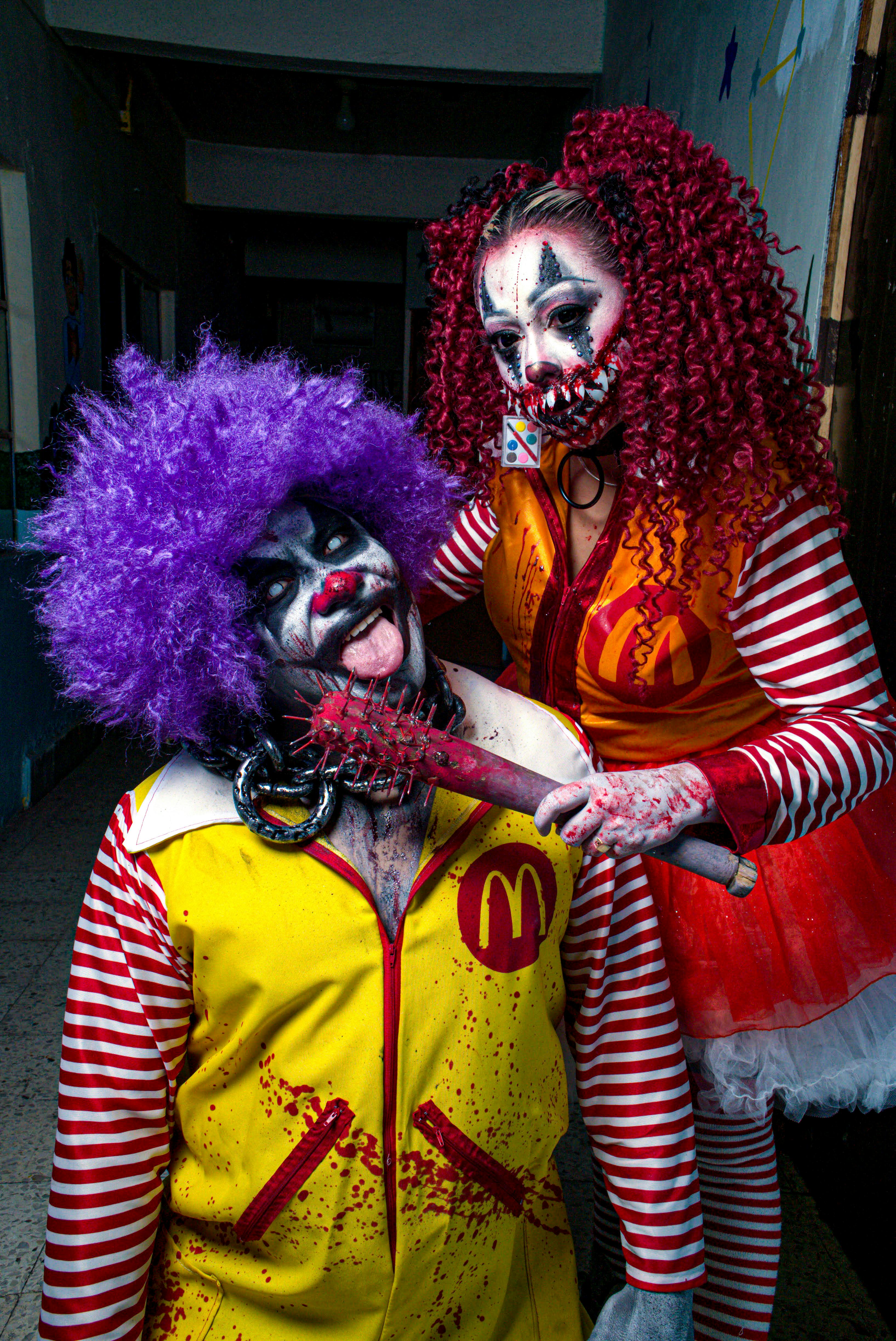 Two adults dressed as sinister, colorful clowns in a dimly lit corridor, perfect for Halloween themes.