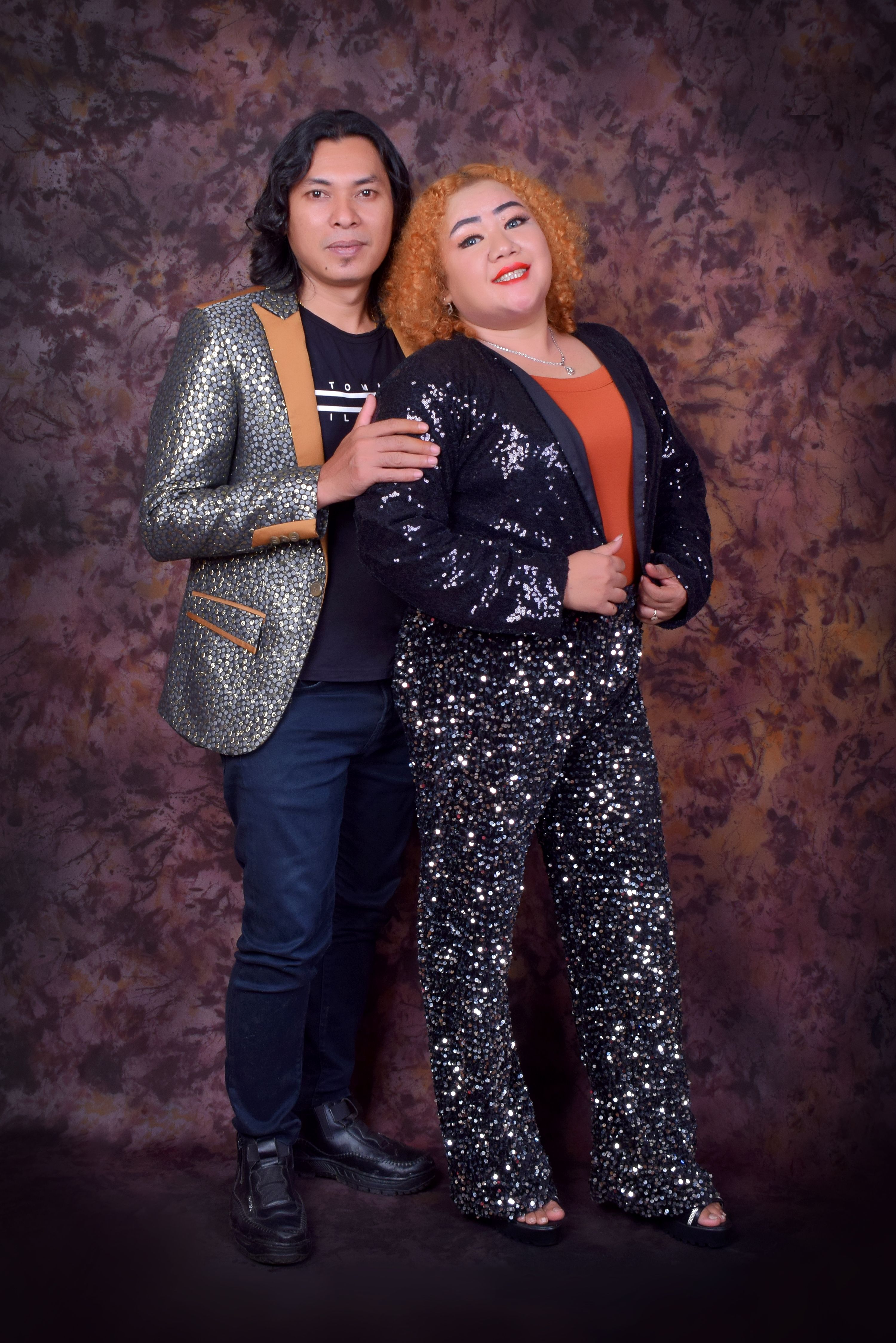 Stylish adult couple posing in glittering outfits against a textured backdrop.