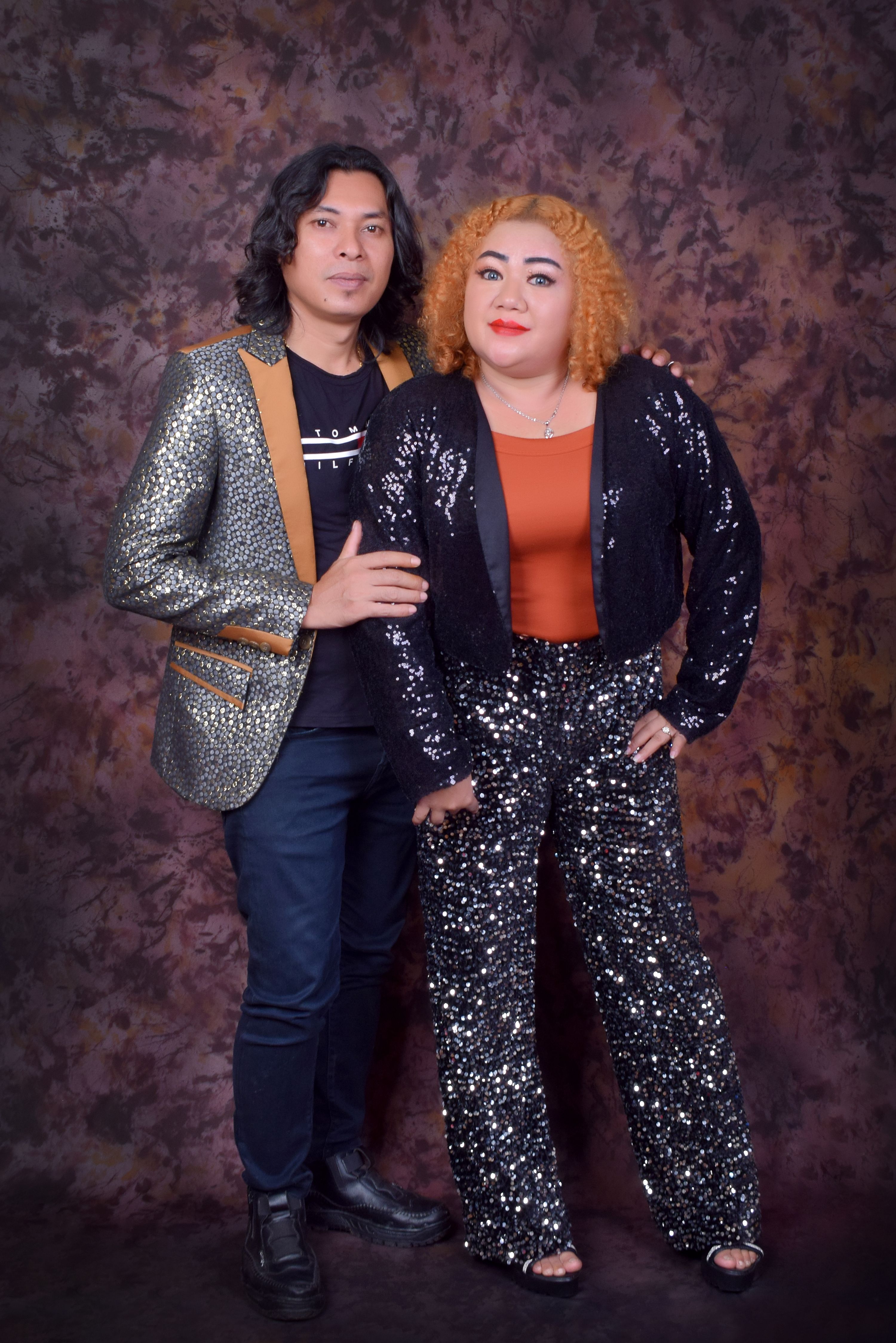 A fashionable couple posing indoors in sparkling sequin attire against a textured backdrop.