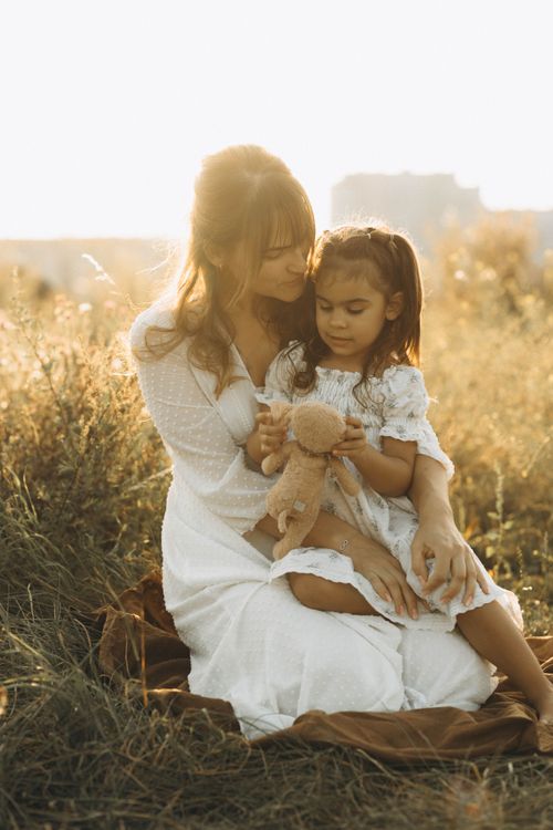 Free Heartwarming moment of mother and daughter embracing in a sunlit field, holding a teddy bear. Stock Photo