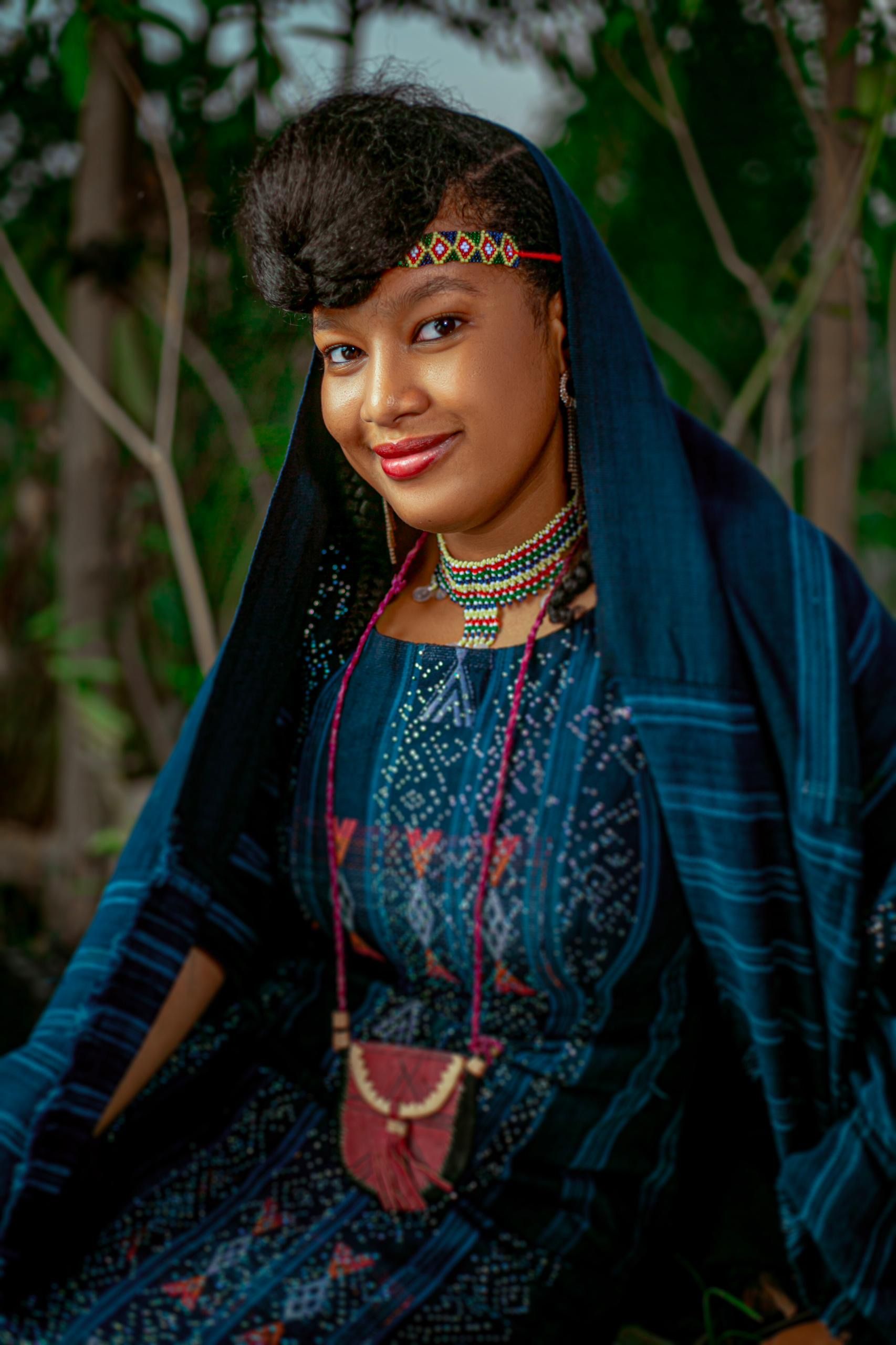 A woman in Nigerian traditional attire poses outdoors, displaying cultural jewelry.