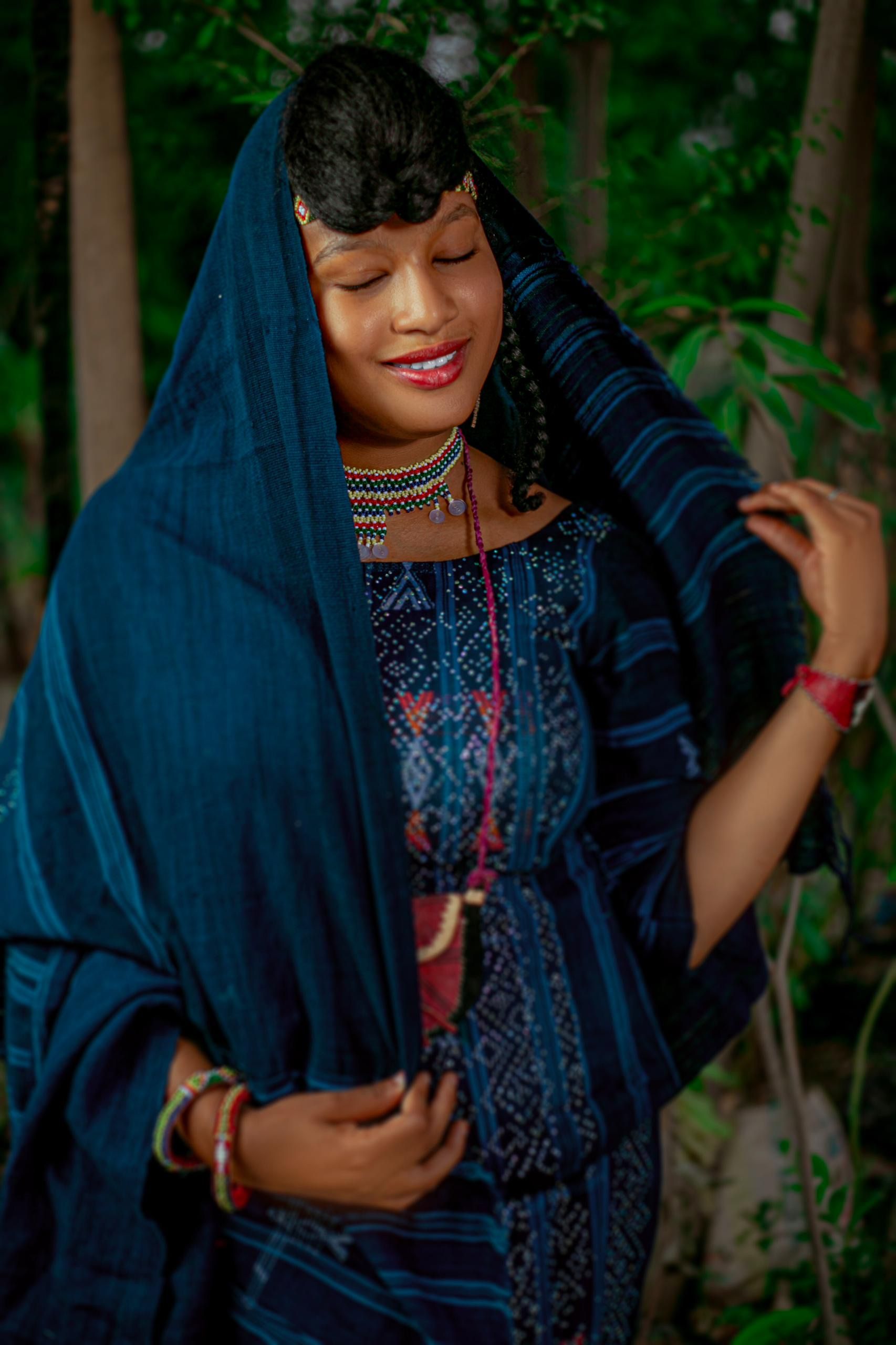 A woman in traditional Nigerian attire smiling gracefully outdoors, surrounded by foliage.
