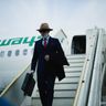 A well-dressed businessman carrying a briefcase steps down the stairs of an airplane onto the tarmac.
