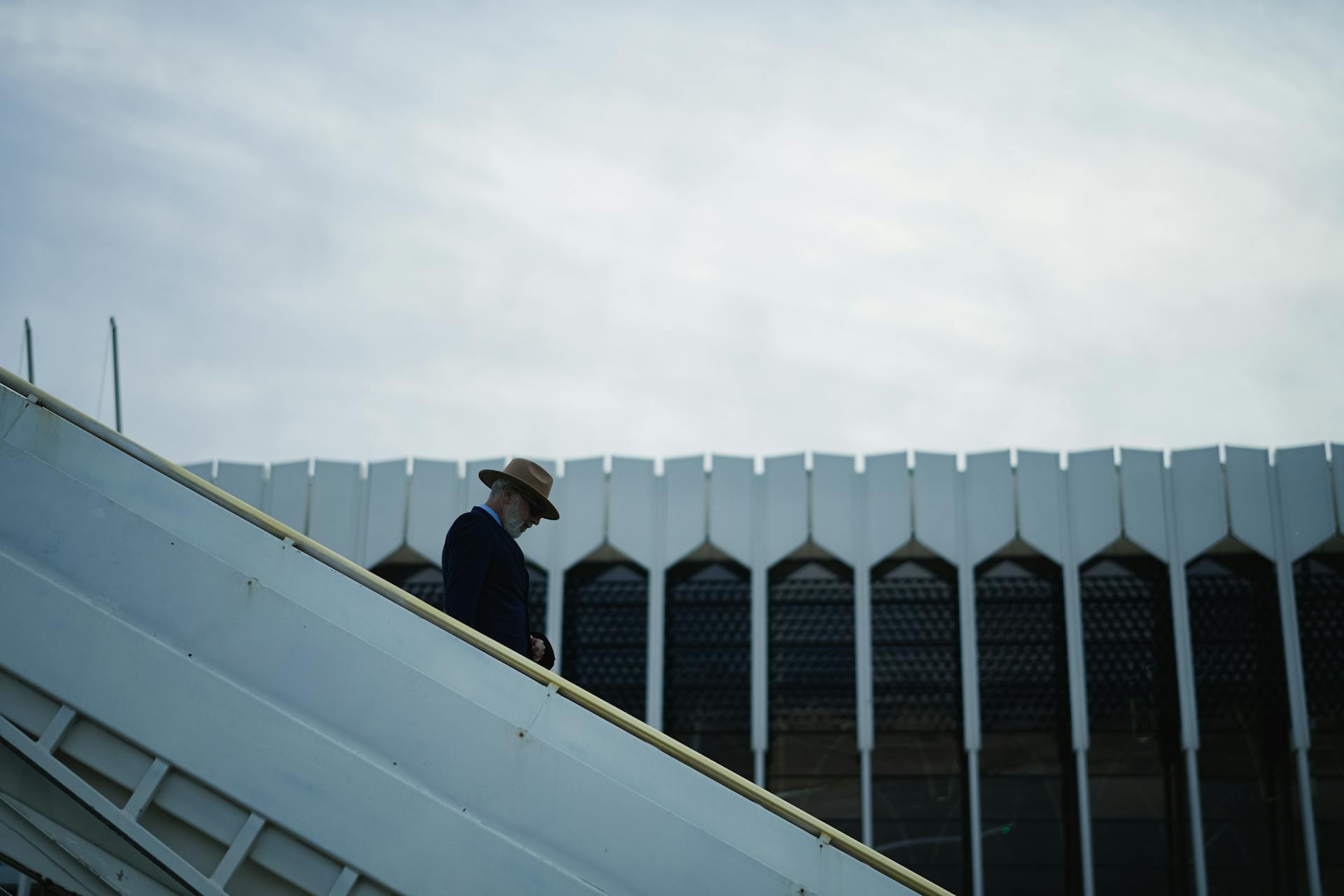 A lone man in a hat ascends airplane stairs with a modern building in the background, emphasizing travel and architecture.