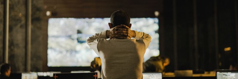 A man stands thoughtfully in a dimly lit control room with various screens.