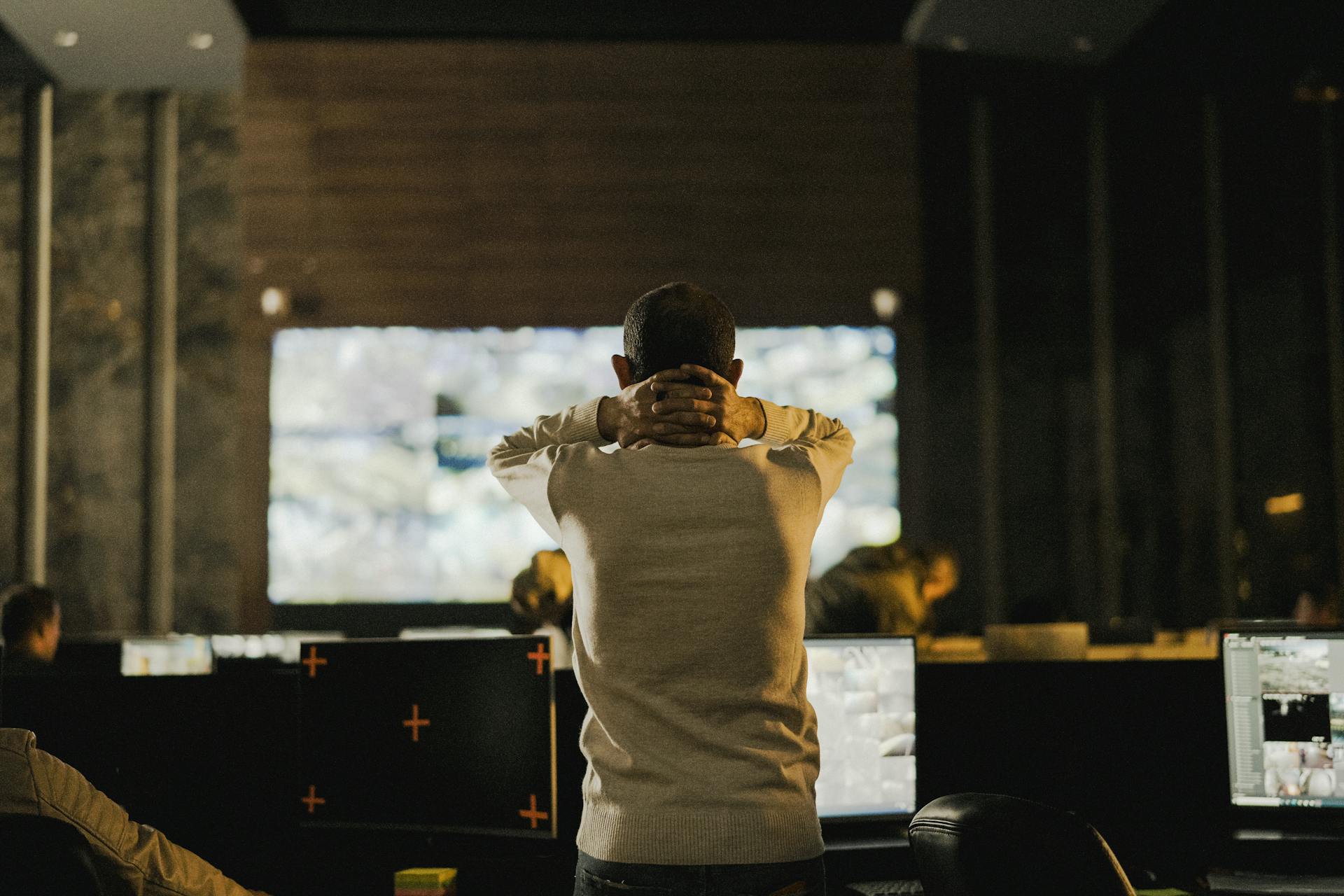 A man stands thoughtfully in a dimly lit control room with various screens.