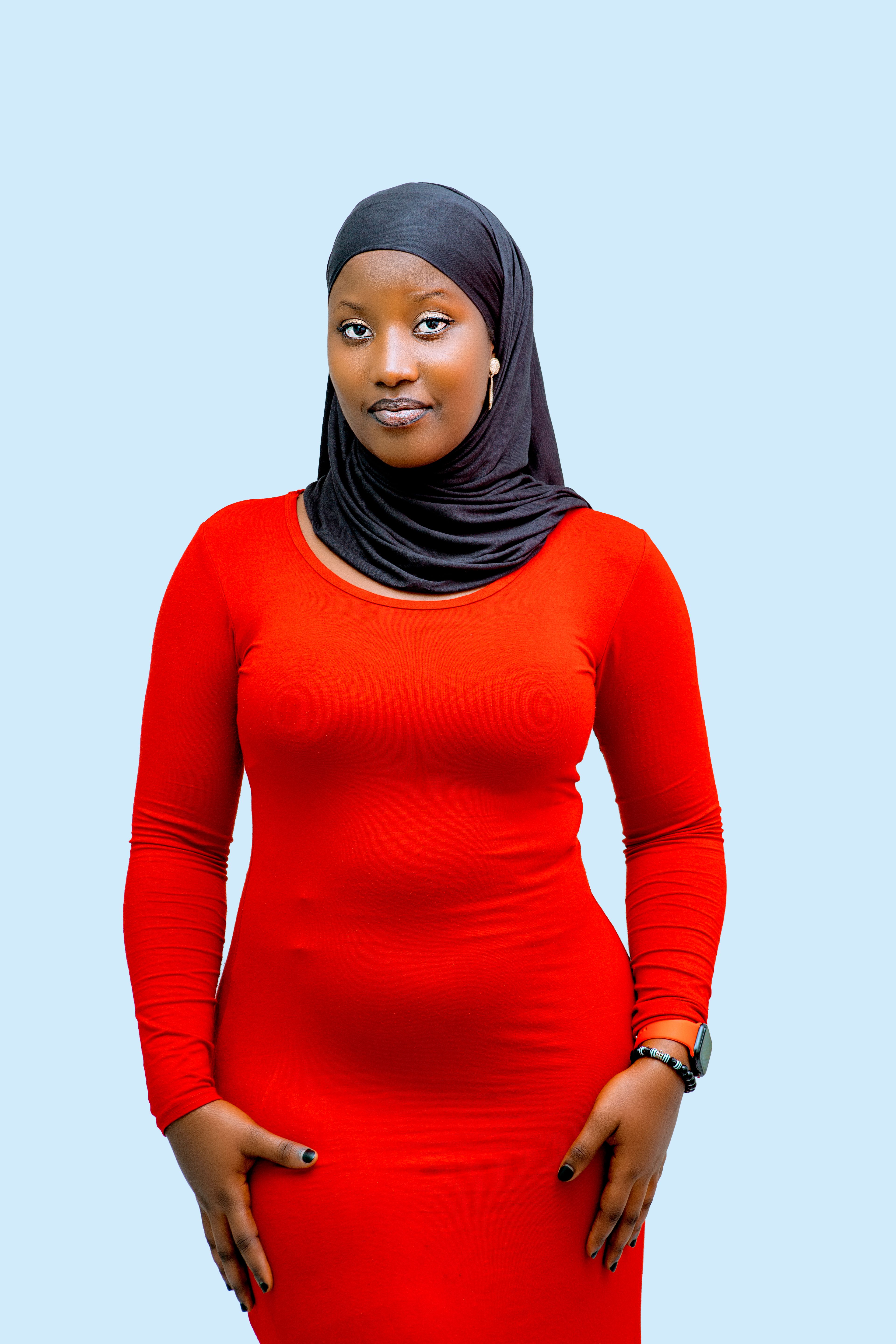 A confident young woman wearing a red dress and hijab standing against a blue background.