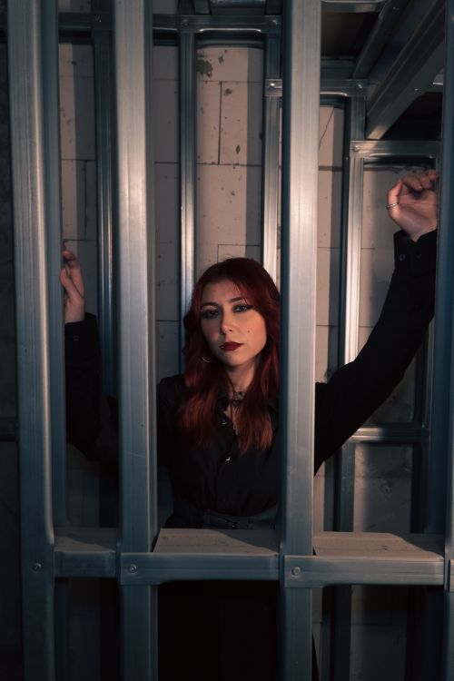Free A young woman with red hair stands behind metal bars in a dimly lit room. Stock Photo