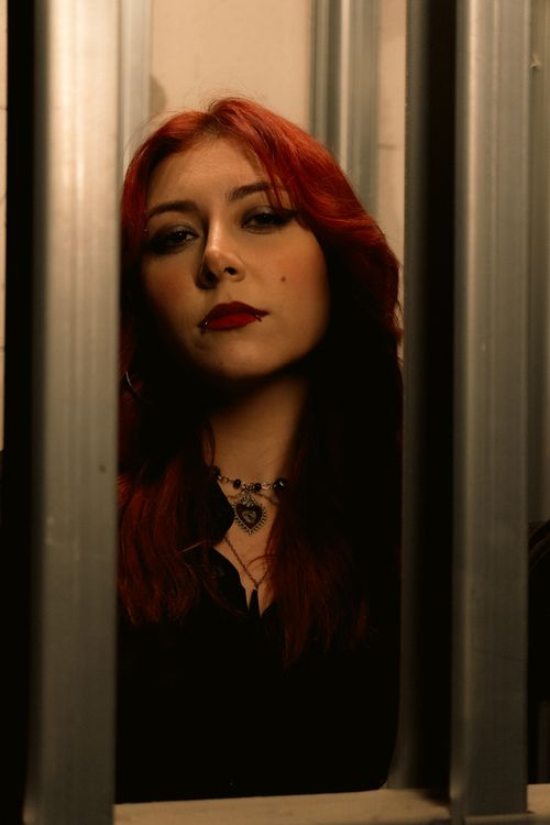 Free Stylish woman with red hair and piercings, framed by metal bars in a dim light setting. Stock Photo