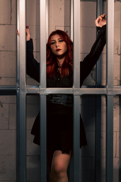 Free Red-haired woman in dark outfit stands behind metal bars, creating a dramatic and mysterious atmosphere. Stock Photo