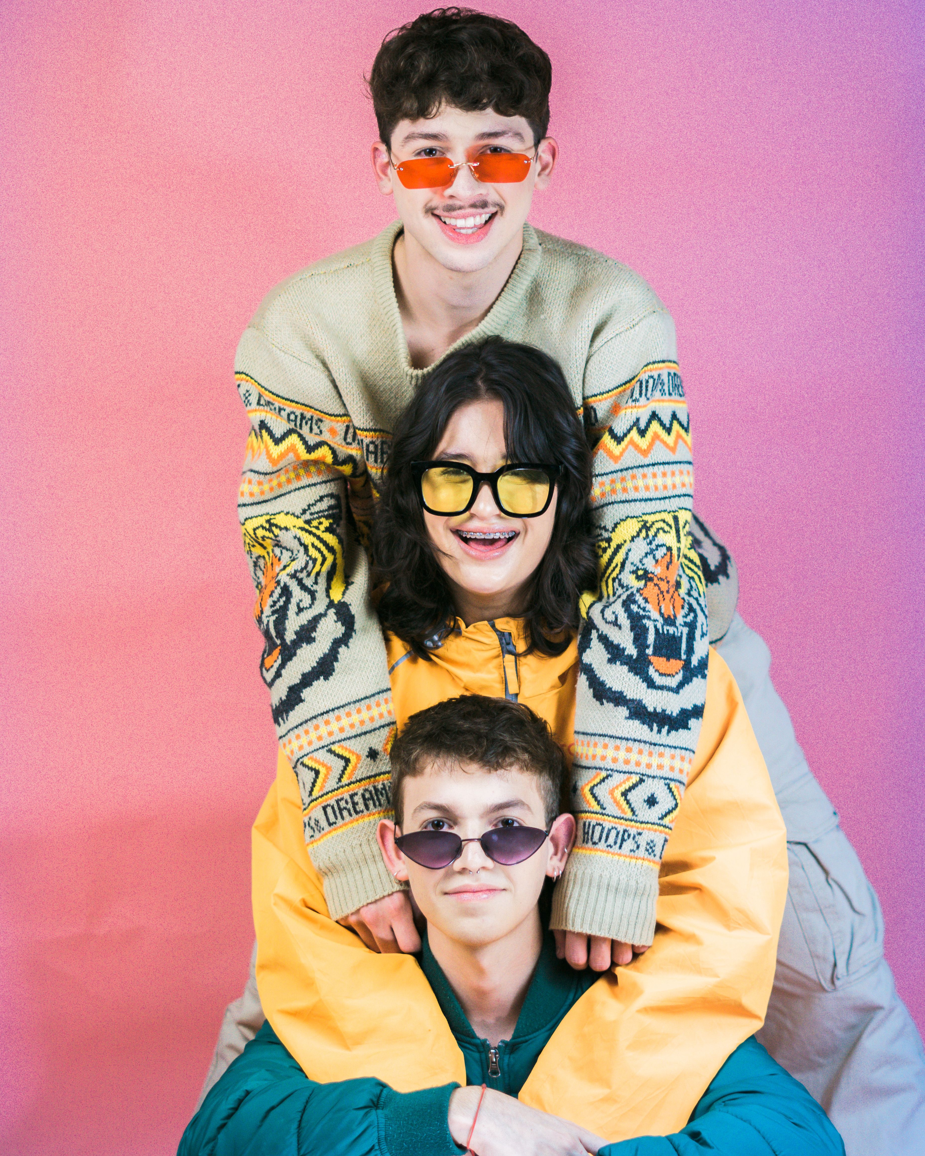 Three stylish teenagers in colorful outfits and sunglasses posing joyfully against a pink backdrop.