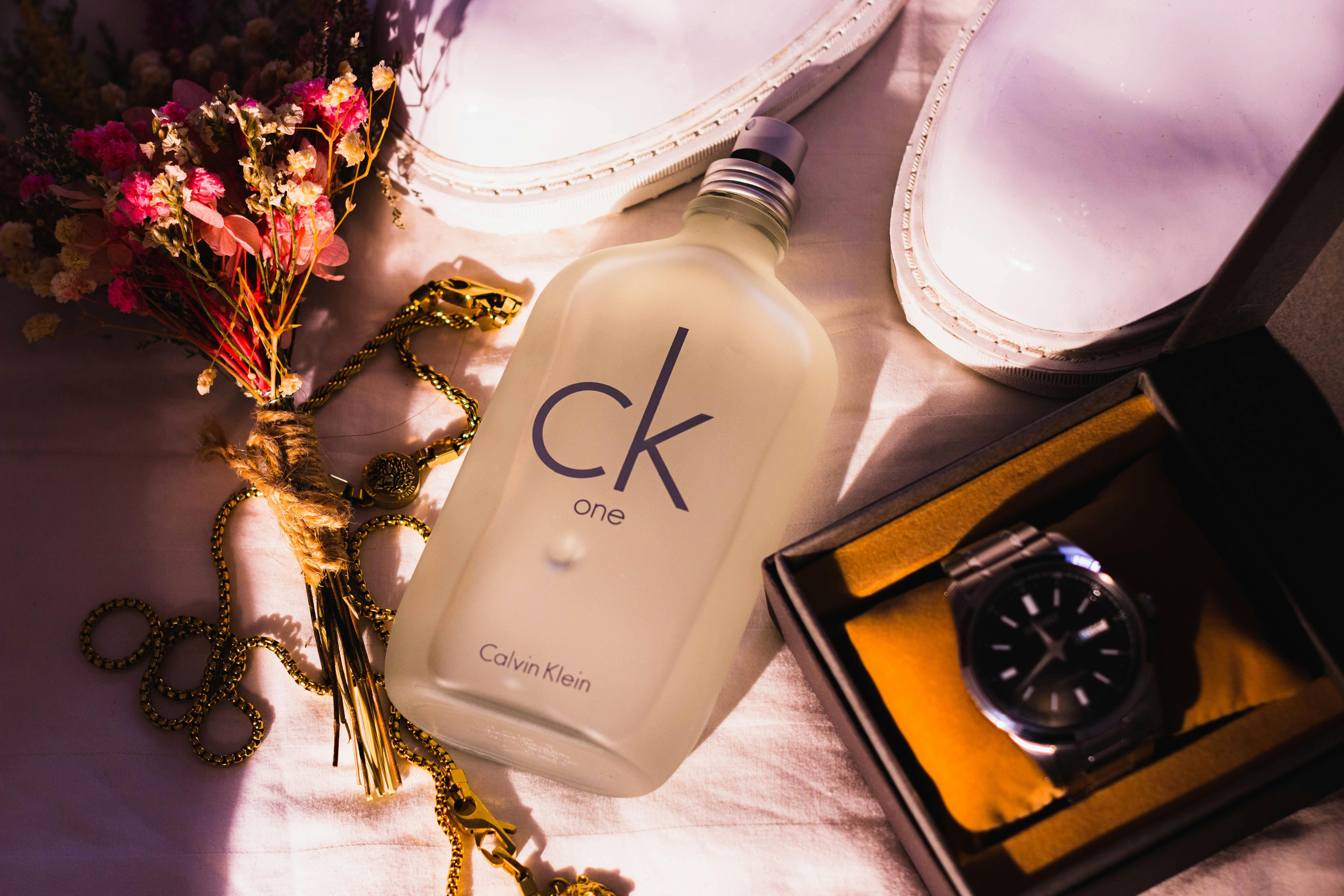 Chic flat lay with CK One bottle, white shoes, watch, flowers and jewelry in warm light.