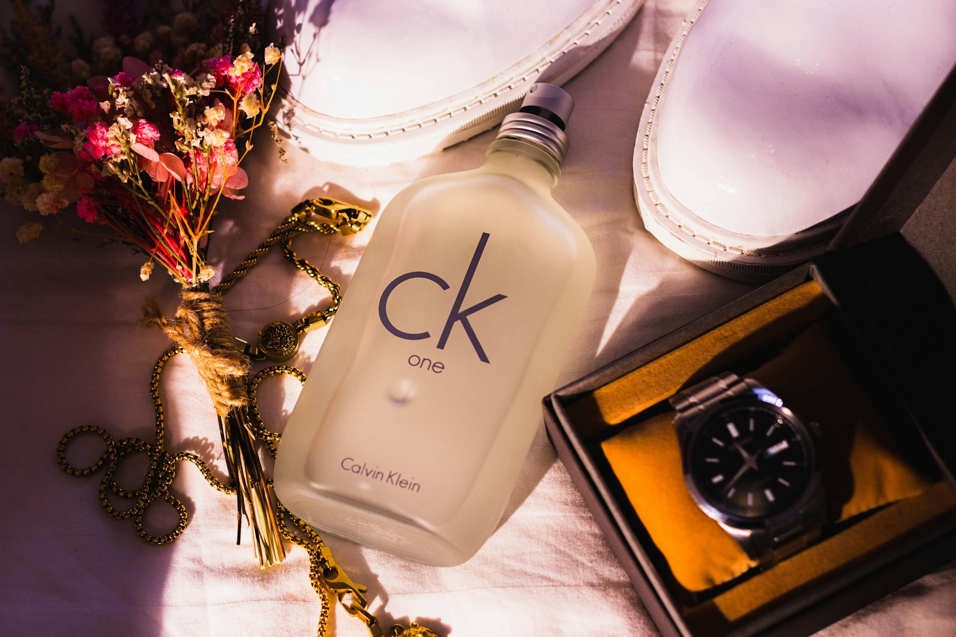 Chic flat lay with CK One bottle, white shoes, watch, flowers and jewelry in warm light.