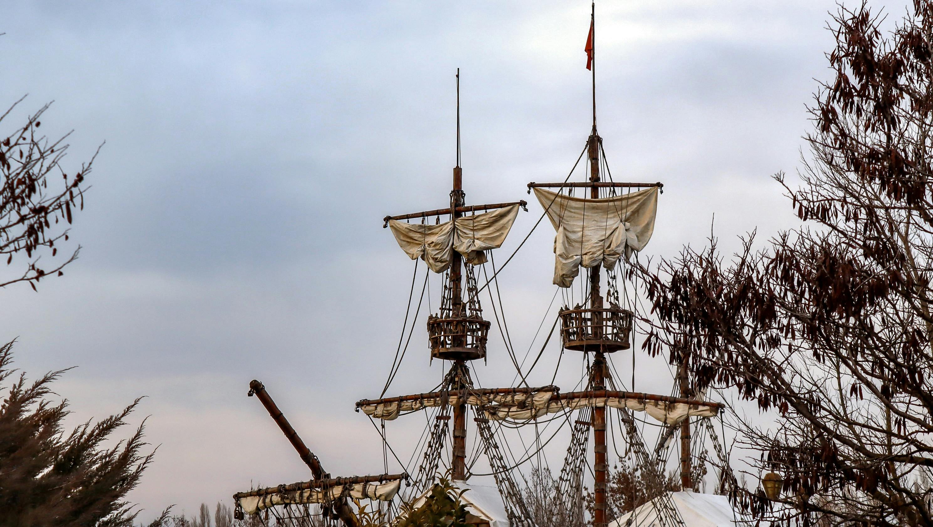 Tall ship masts with tattered sails amidst bare trees under a cloudy sky.