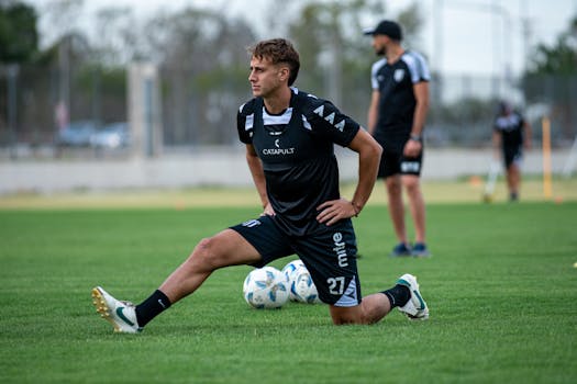 A soccer player stretches on the field during training, surrounded by teammates and soccer balls.