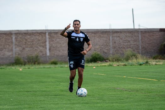 Soccer player in black jersey dribbling a ball on a grassy field during daytime training session.