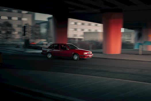 A red vintage car speeds through a moody urban landscape in Scotland, showcasing dynamic motion and architectural contrast.