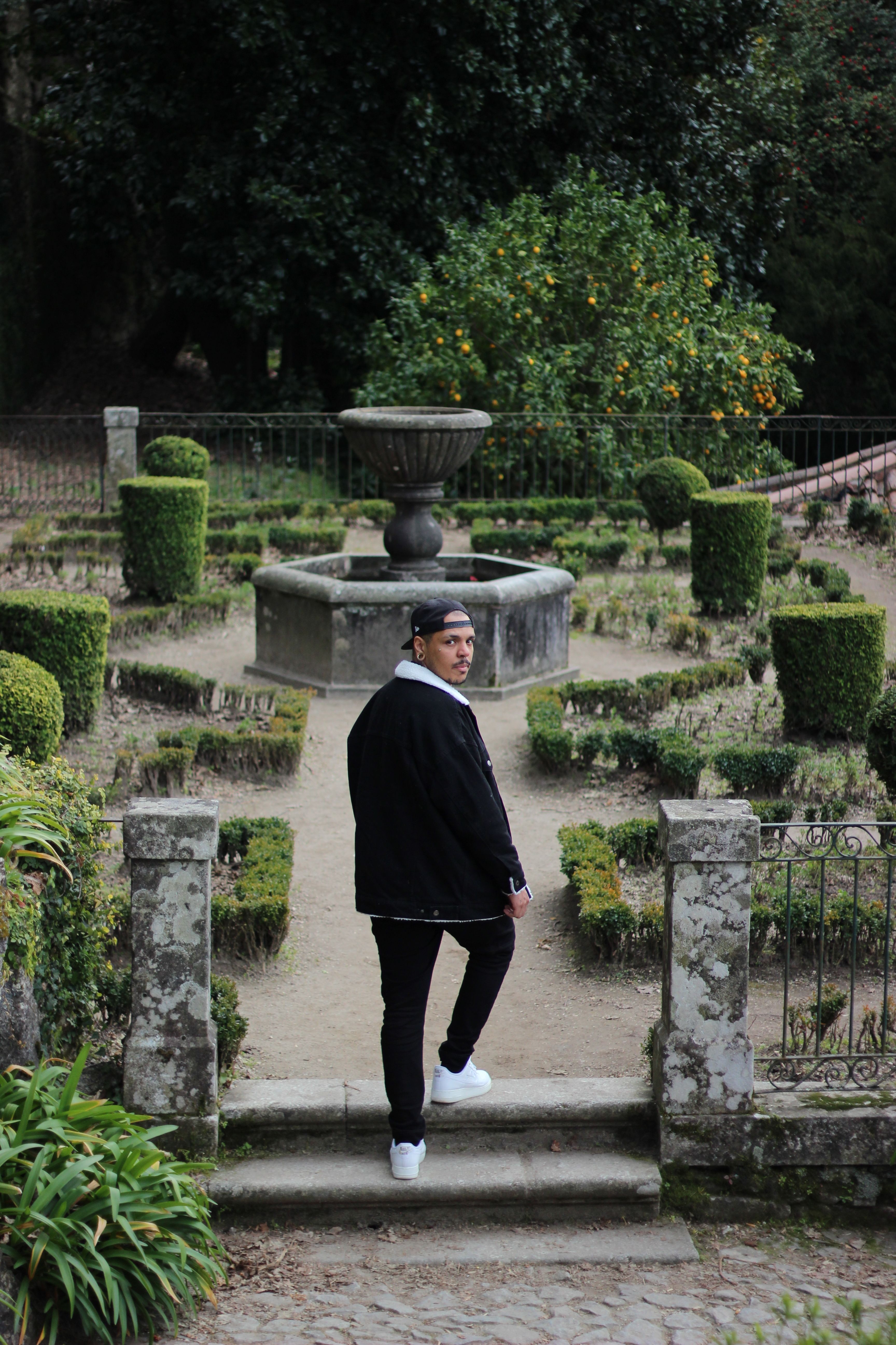 A stylish man in a coat walks through a classic garden with a fountain centerpiece.