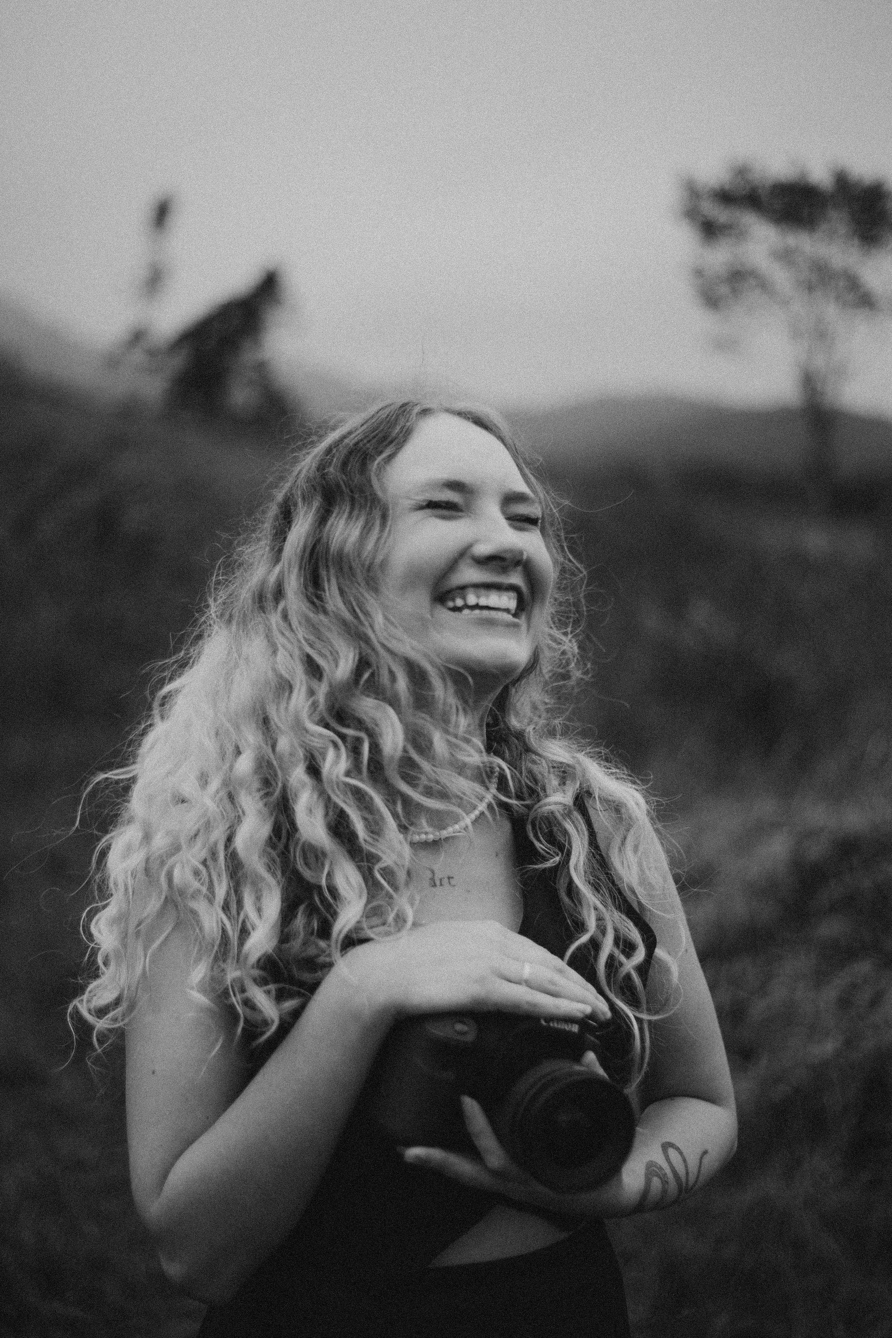 Black and white portrait of a young woman holding a camera and smiling in a natural setting.