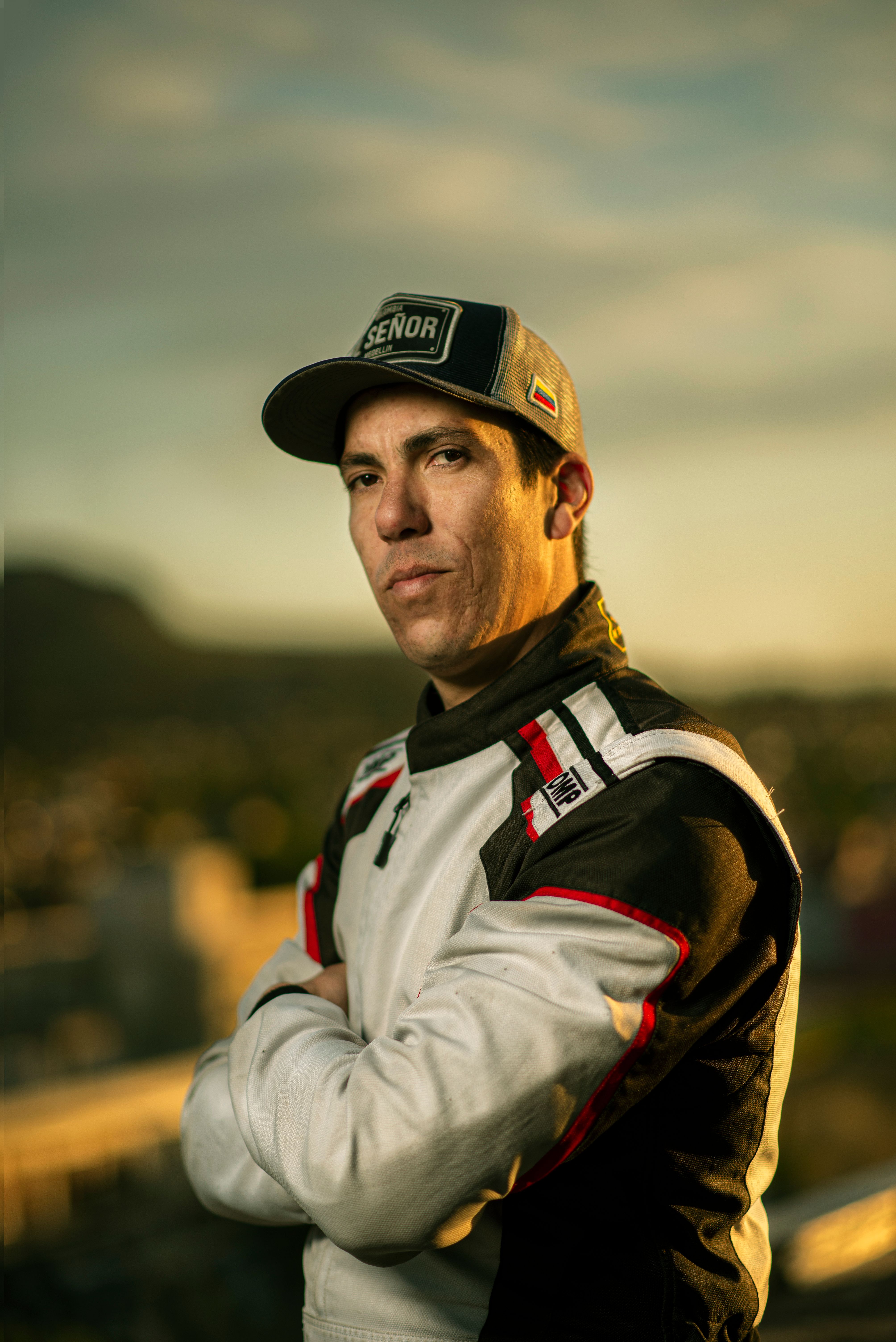Confident race car driver posing outdoors in racing suit with sunset backdrop.