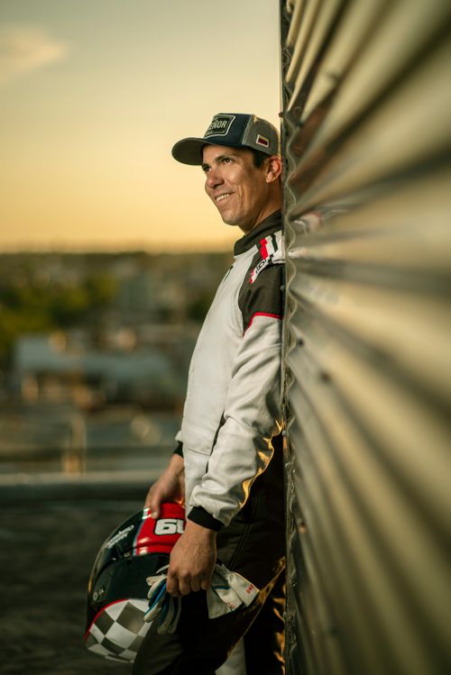 Free A professional race car driver in uniform standing outdoors at sunset, holding a helmet. Stock Photo