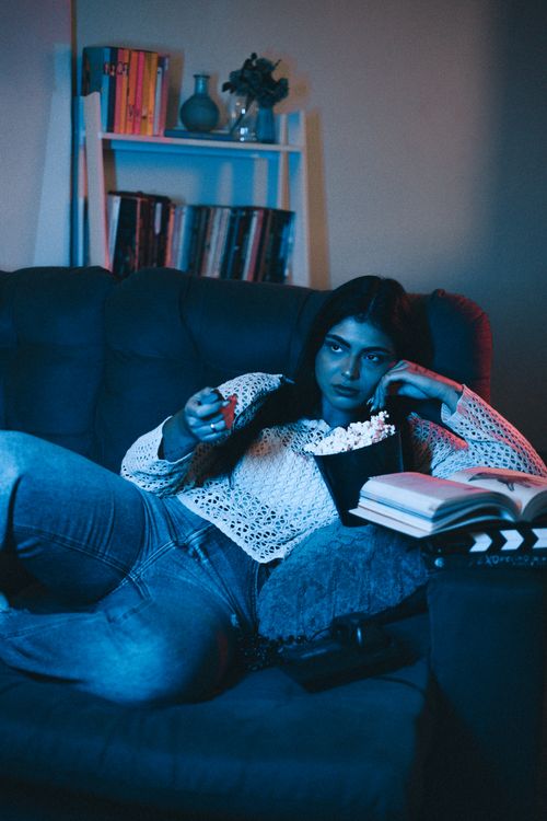Free Young woman enjoying a cozy night in, watching TV with popcorn and books. Stock Photo