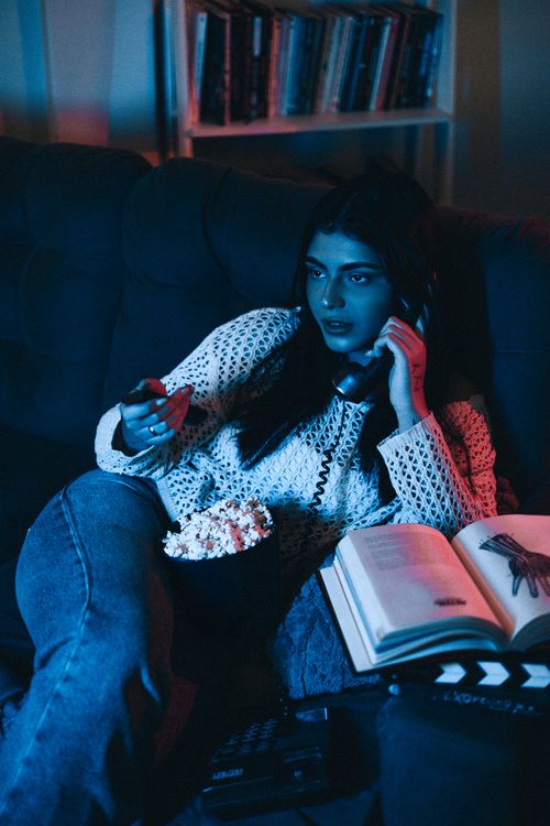 Free Young woman watching TV with remote, popcorn, and headphones in a cozy room setting. Stock Photo