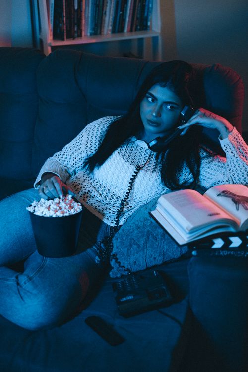Free Woman relaxing on a sofa with popcorn, an open book, and a vintage phone in a cozy home setting. Stock Photo