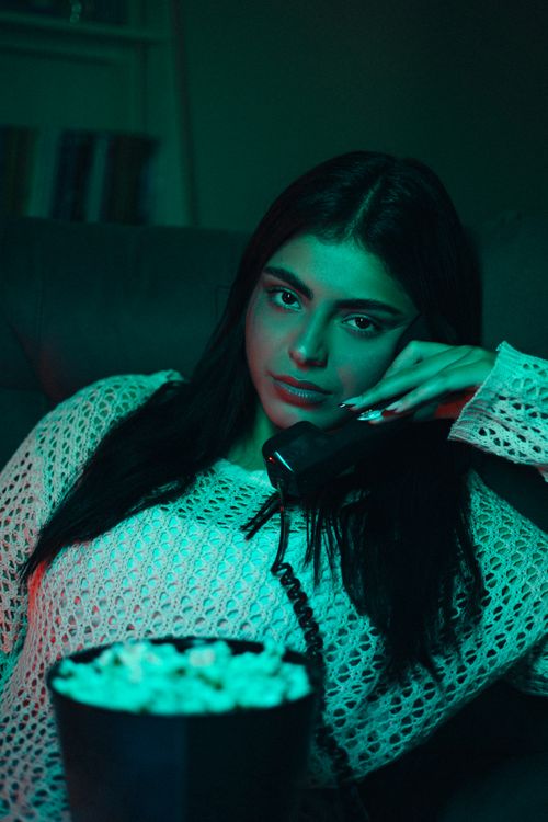 Free Young woman relaxing indoors, holding phone, with bowl of popcorn, ambient lighting. Stock Photo