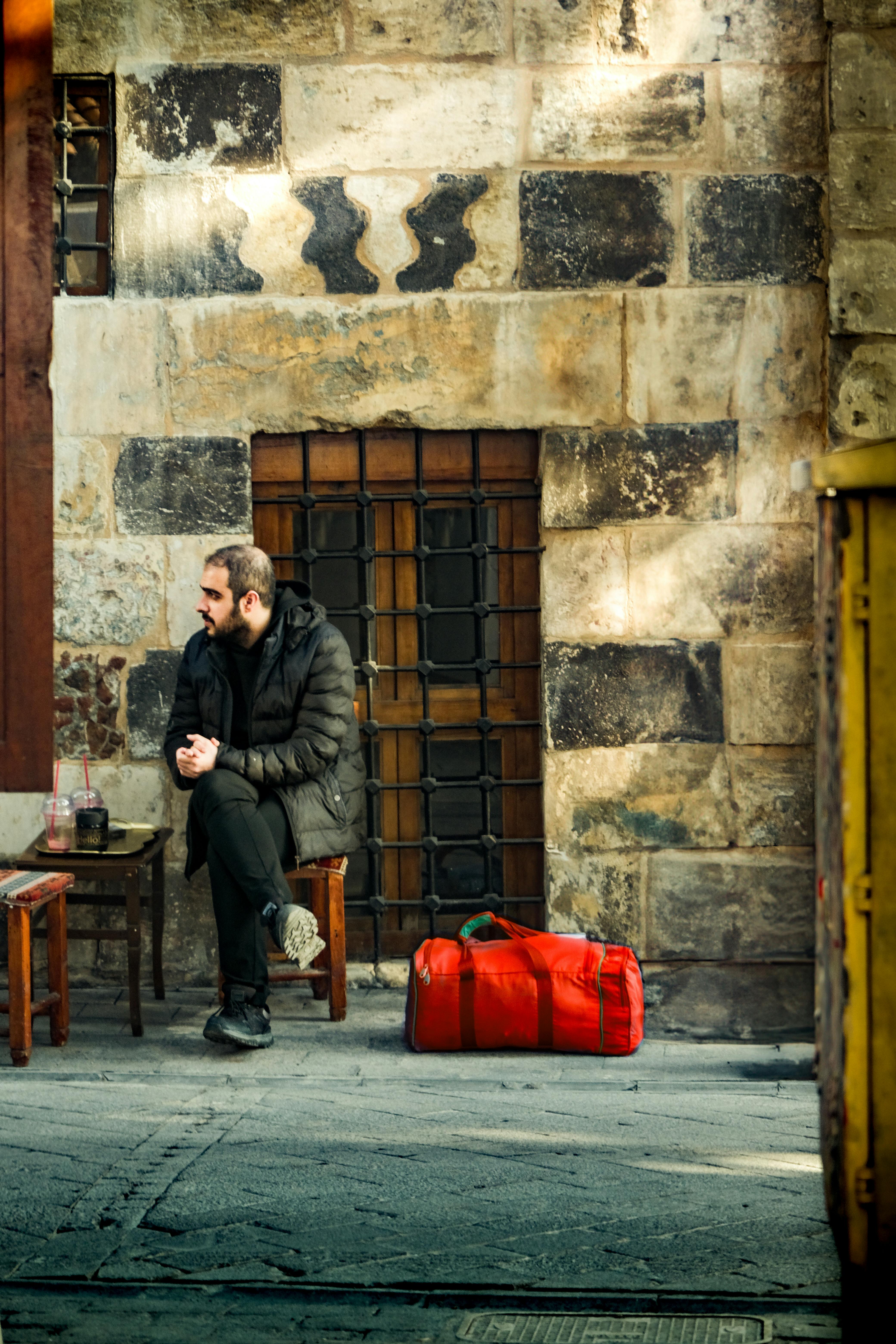 A man sits beside a red bag by a rustic stone wall in daylight.
